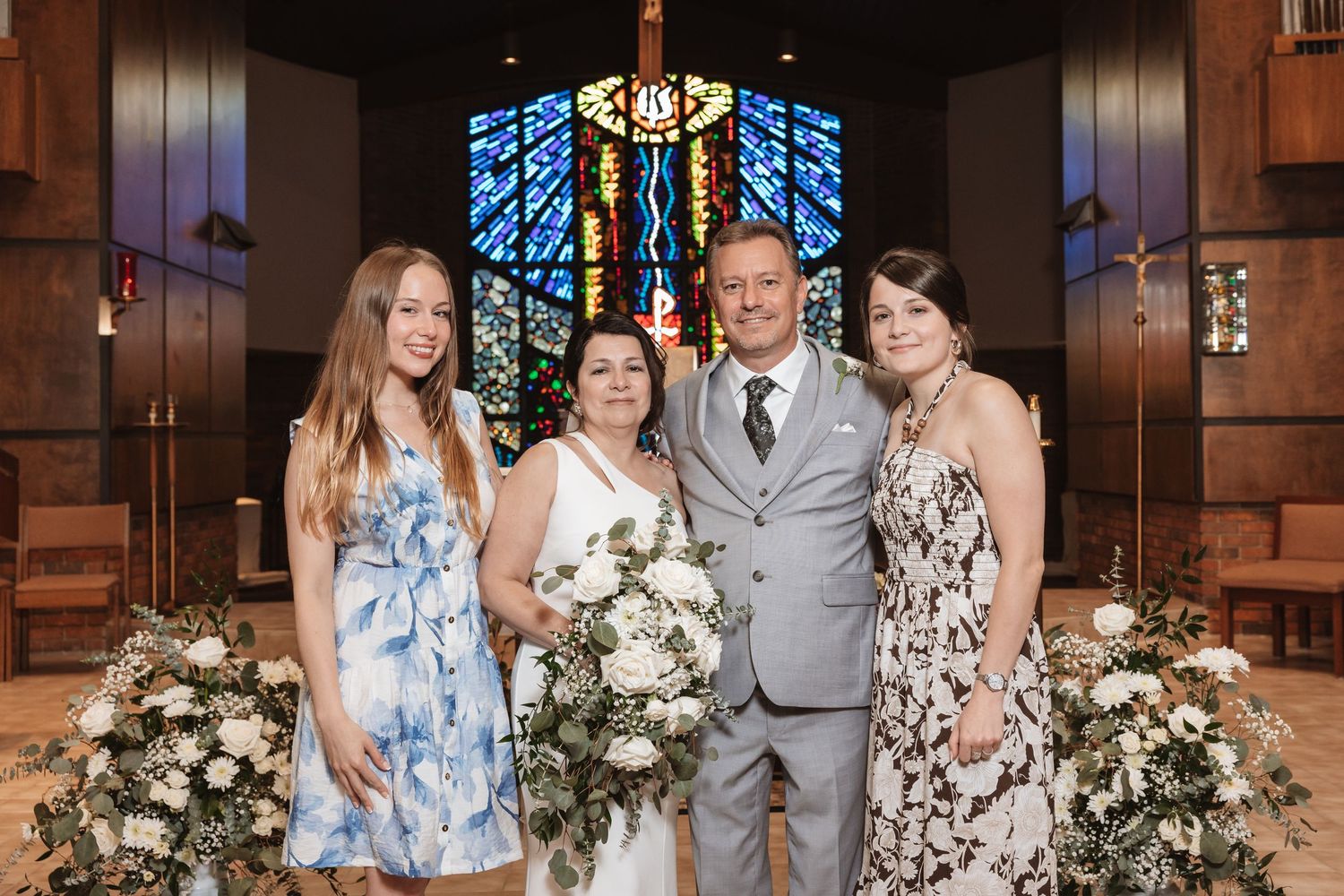 Bride and groom pose with two daughters in front of stained glass altar at church wedding ceremony.
