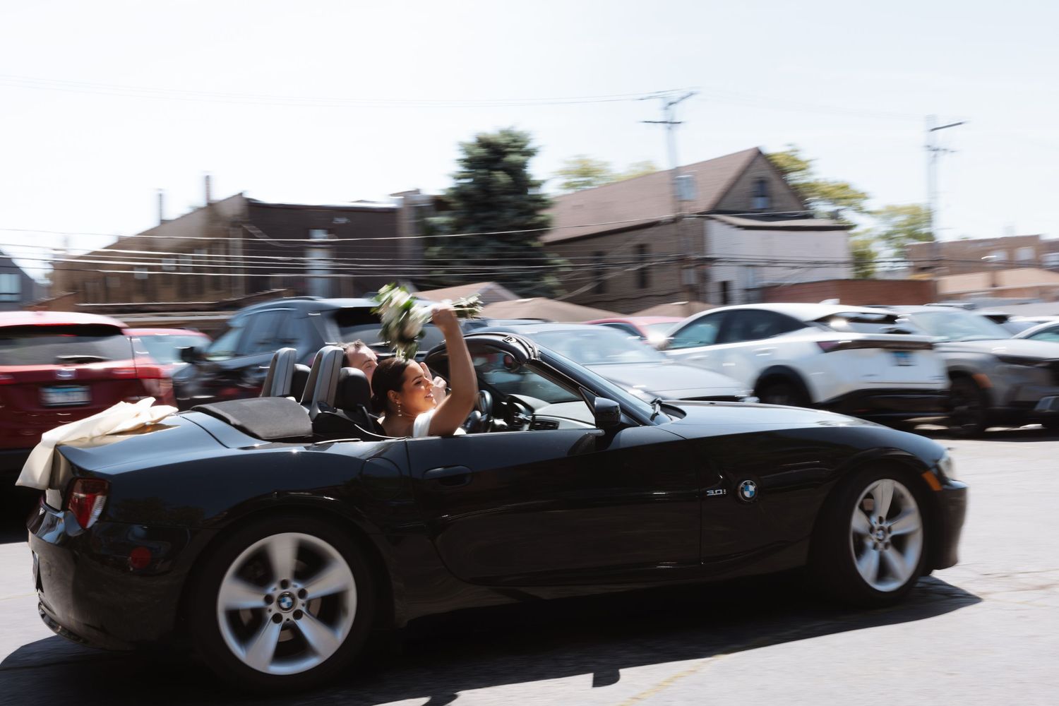 Newlyweds celebrate in black BMW convertible with bouquet raised in sunny parking lot.