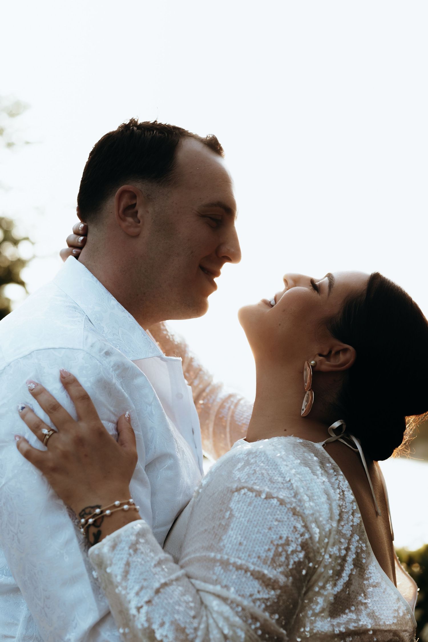 Bride and groom share romantic moment in white sequin outfits with soft backlit glow outdoors.