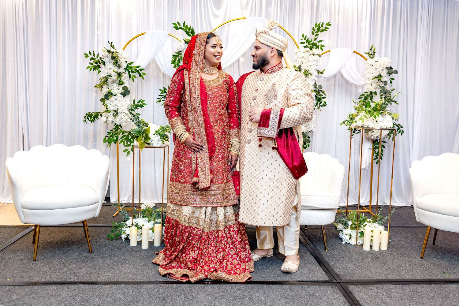 South Asian wedding couple in traditional red and gold attire standing together before elegant white backdrop.