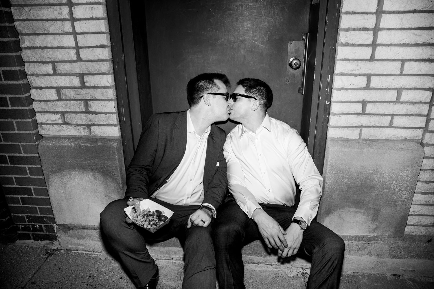 Black and white photo of two grooms kissing on steps outside a brick building at wedding.