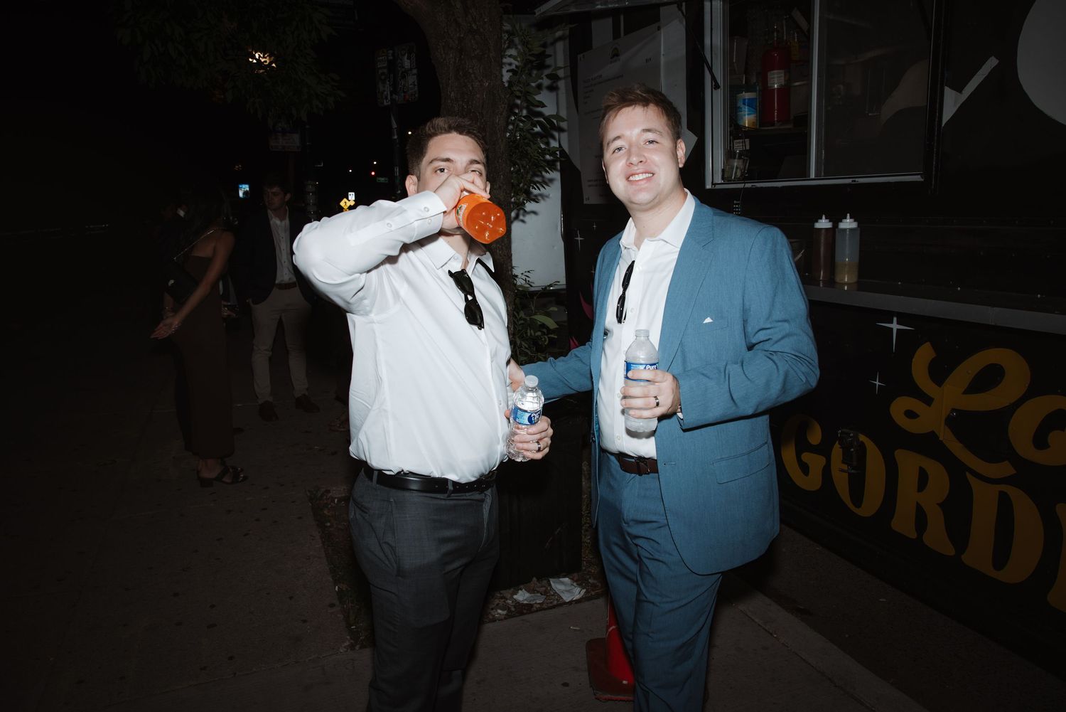 Two men in suits smile and drink beverages outside a food truck at night.