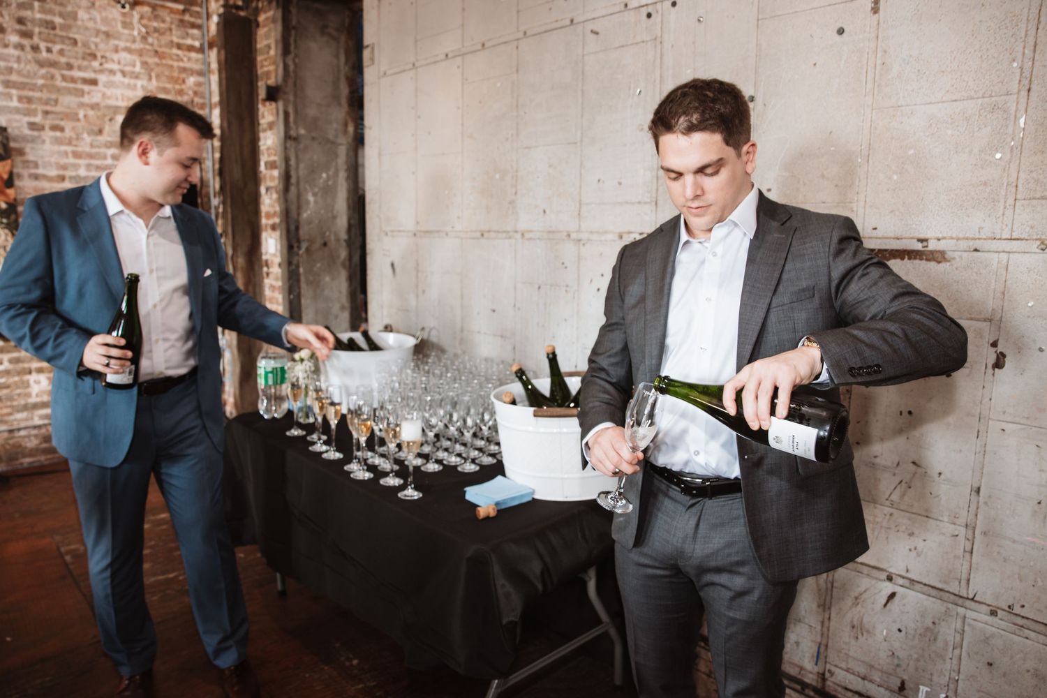 Two men in suits pour champagne into glasses at a beverage station during an event.