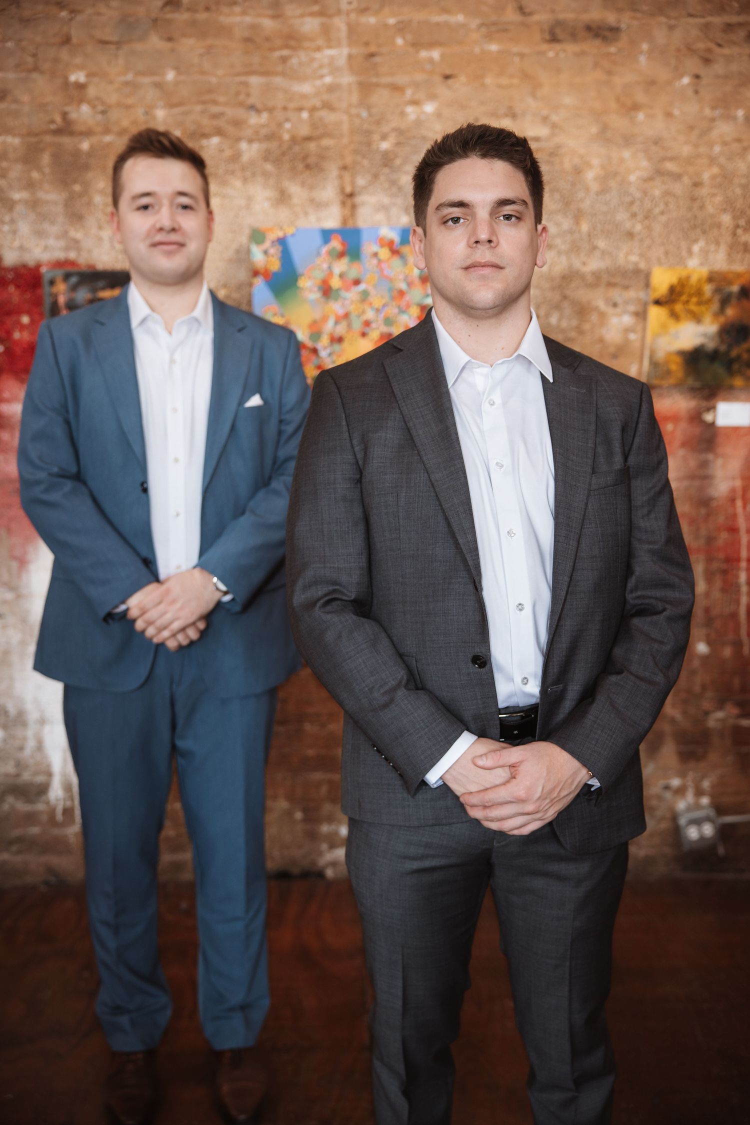 Two young men in suits pose formally in an art gallery with brick walls and colorful paintings.