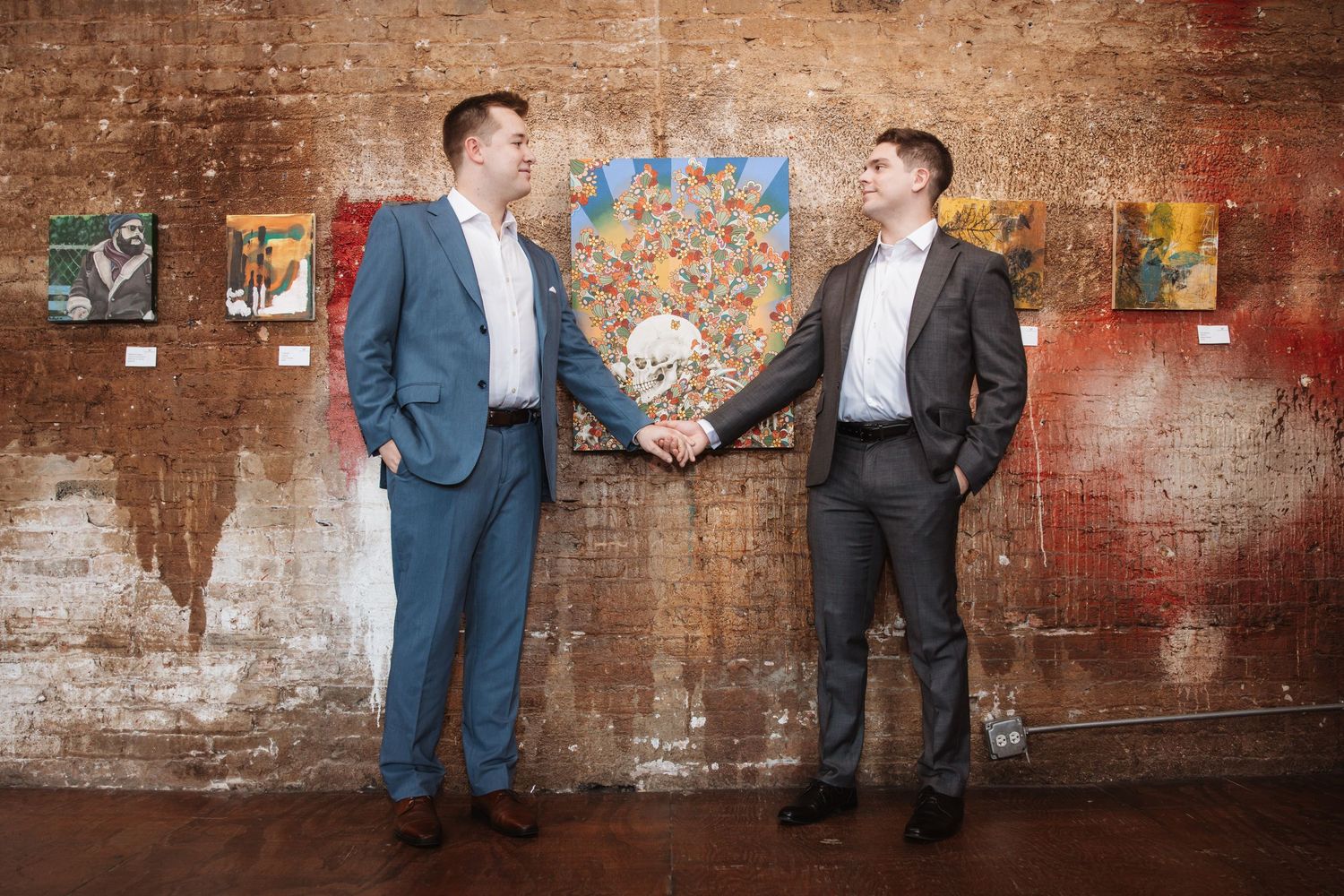 Two men in suits shaking hands and smiling in front of colorful artwork on a brick wall.