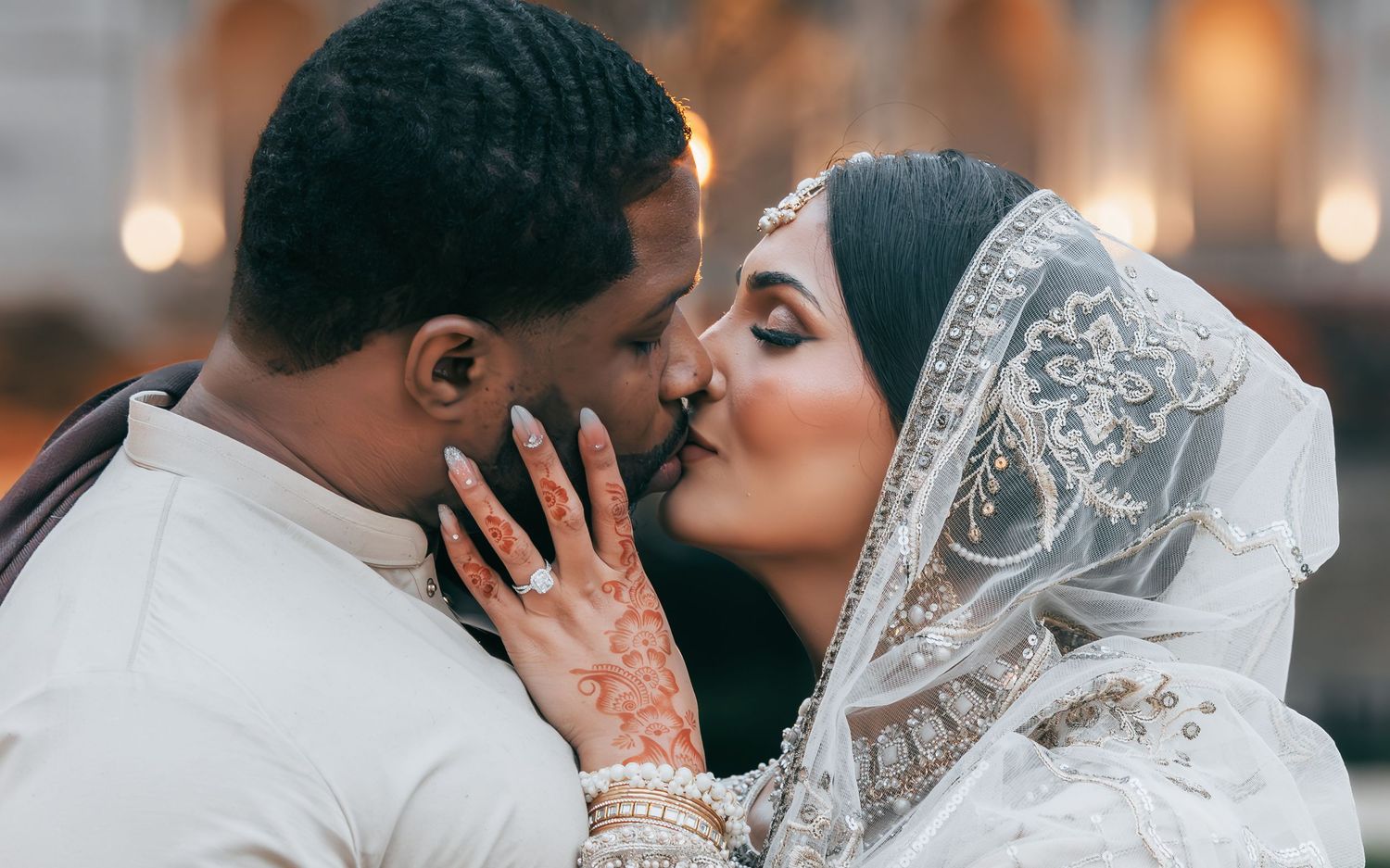 Interracial Muslim couple sharing tender kiss, bride wearing ornate ivory lehenga with lace dupatta veil.