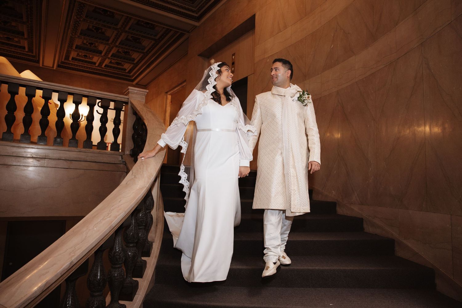 Bride and groom in wedding attire walk hand in hand down an elegant wooden staircase.