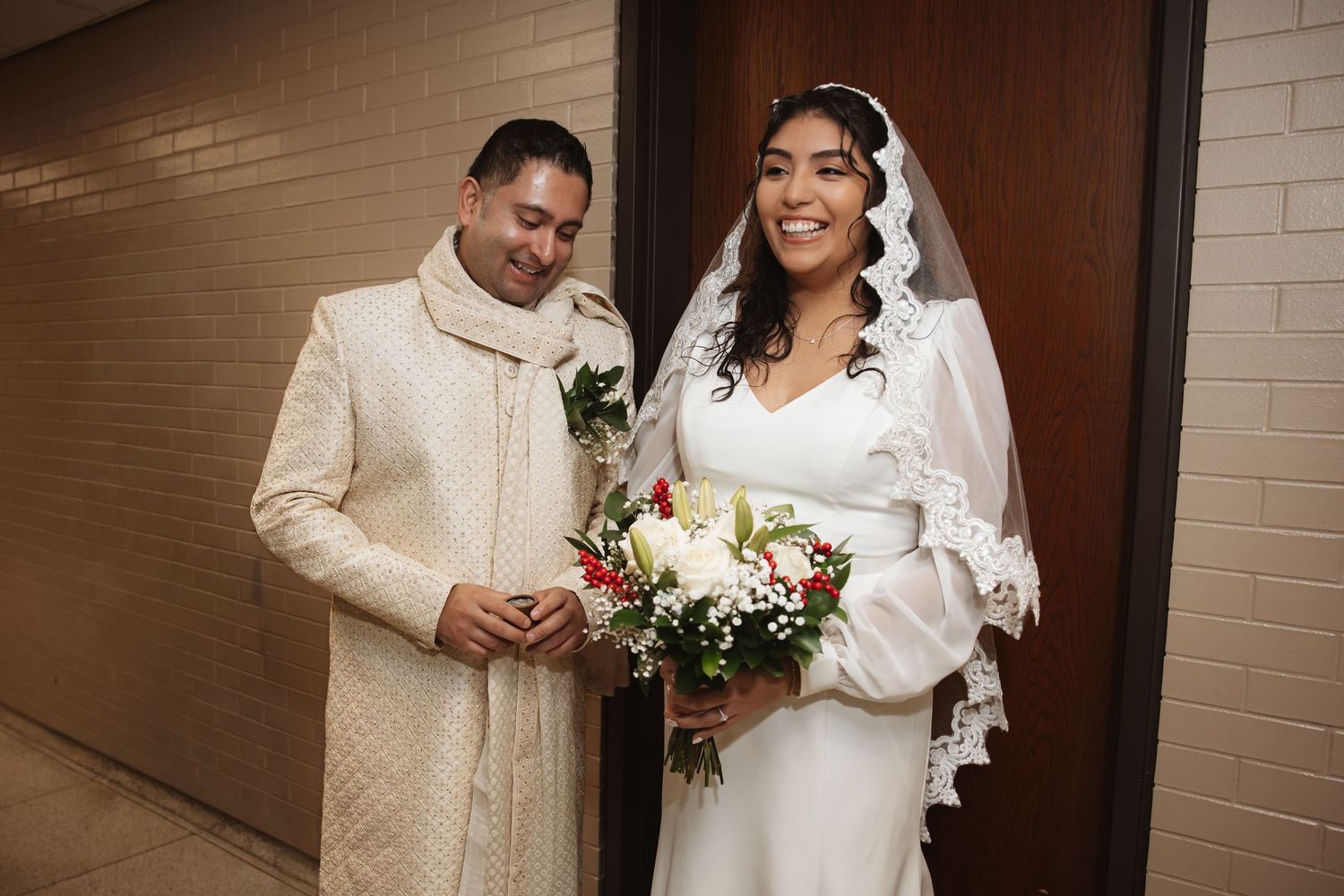 Smiling bride in white lace veil holds floral bouquet while standing beside groom in ivory embroidered sherwani.