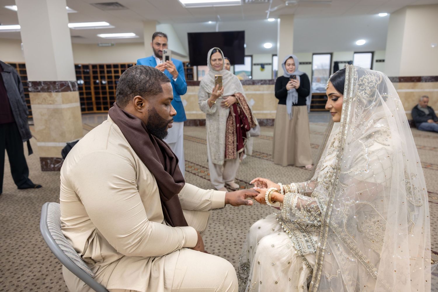 Groom placing ring on bride's finger during Islamic nikah ceremony inside a mosque with guests watching.