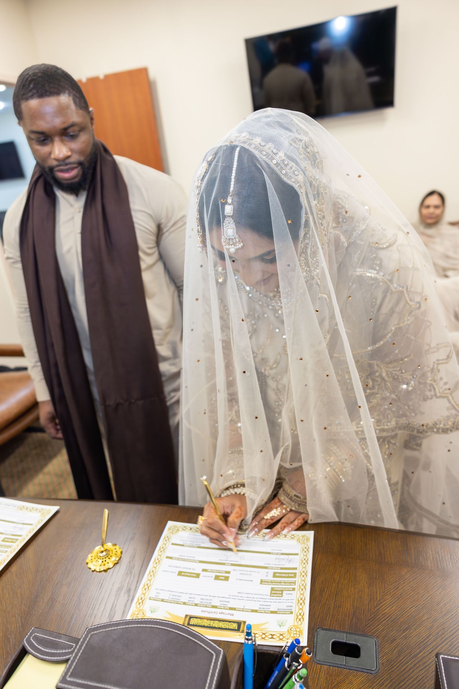 South Asian bride in white embroidered lehenga signing nikah marriage contract at Islamic wedding ceremony.