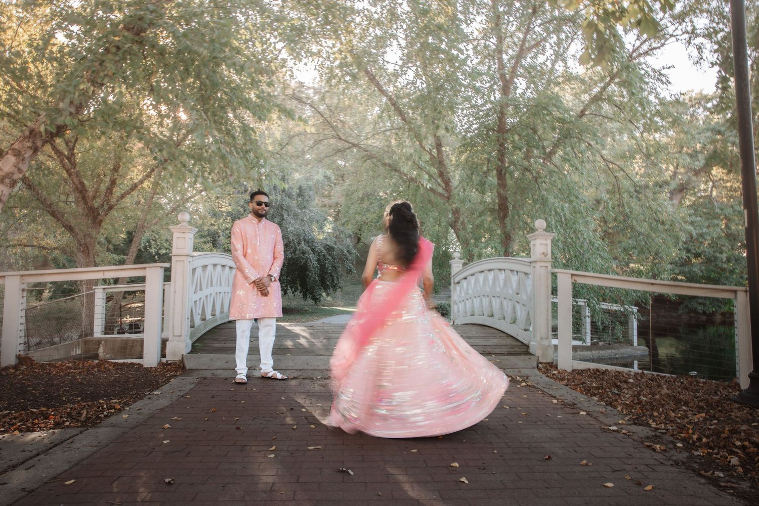 Bride and groom in coordinating pink attire on tree-lined path by white bridge.