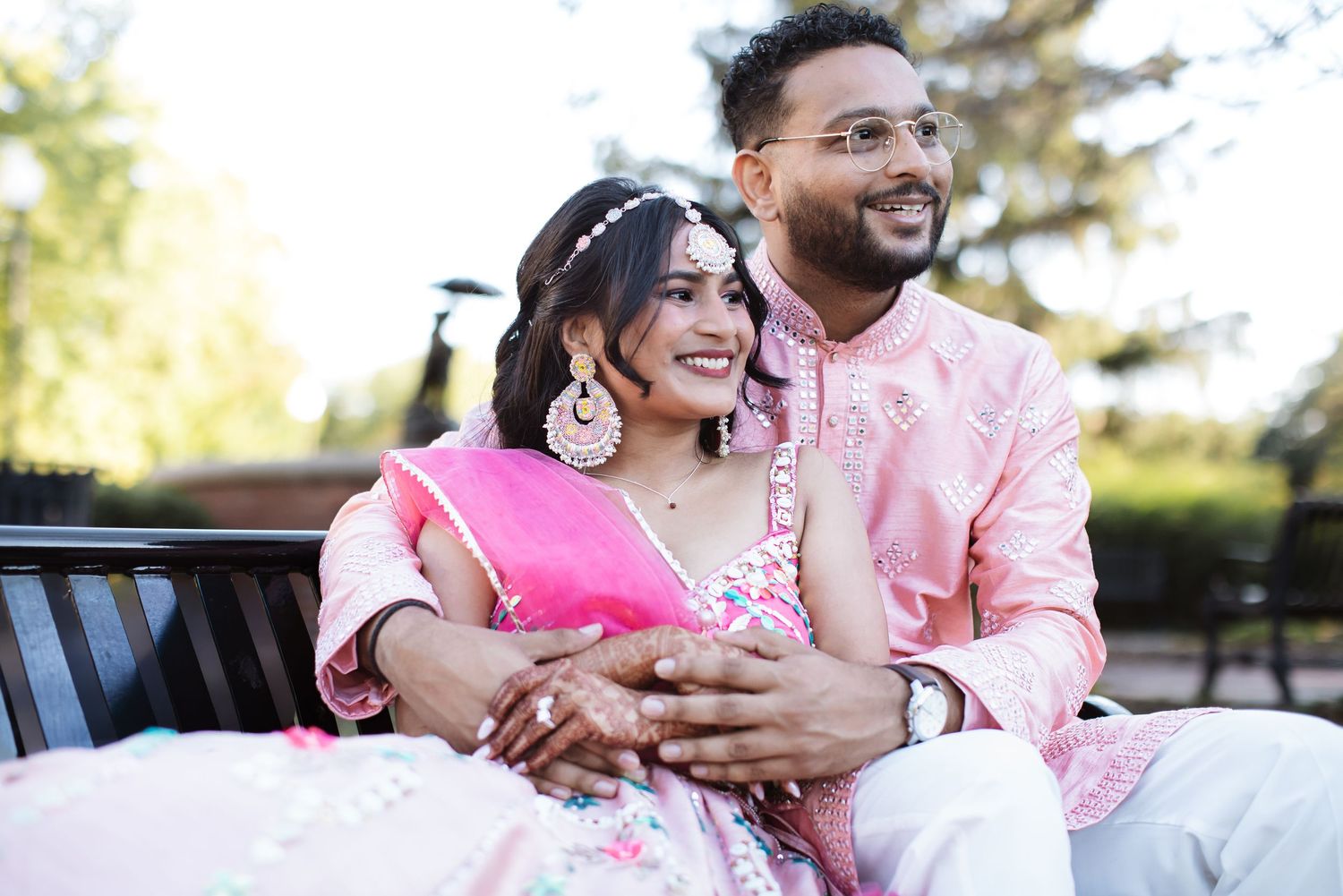 Indian couple in pink traditional wedding outfits sitting on a bench gazing upward.