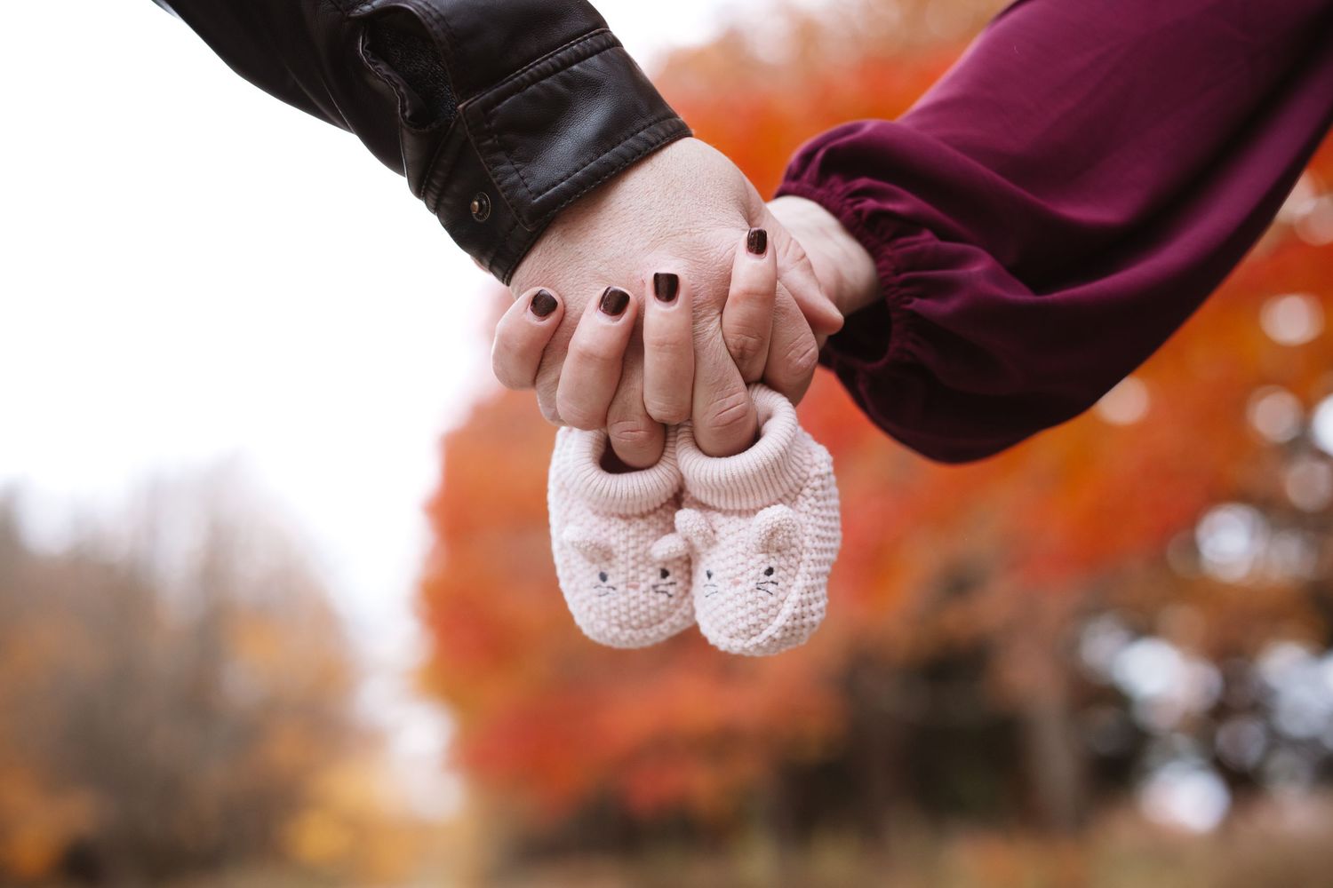 Couple holding hands and tiny baby booties in autumn, pregnancy announcement photo.