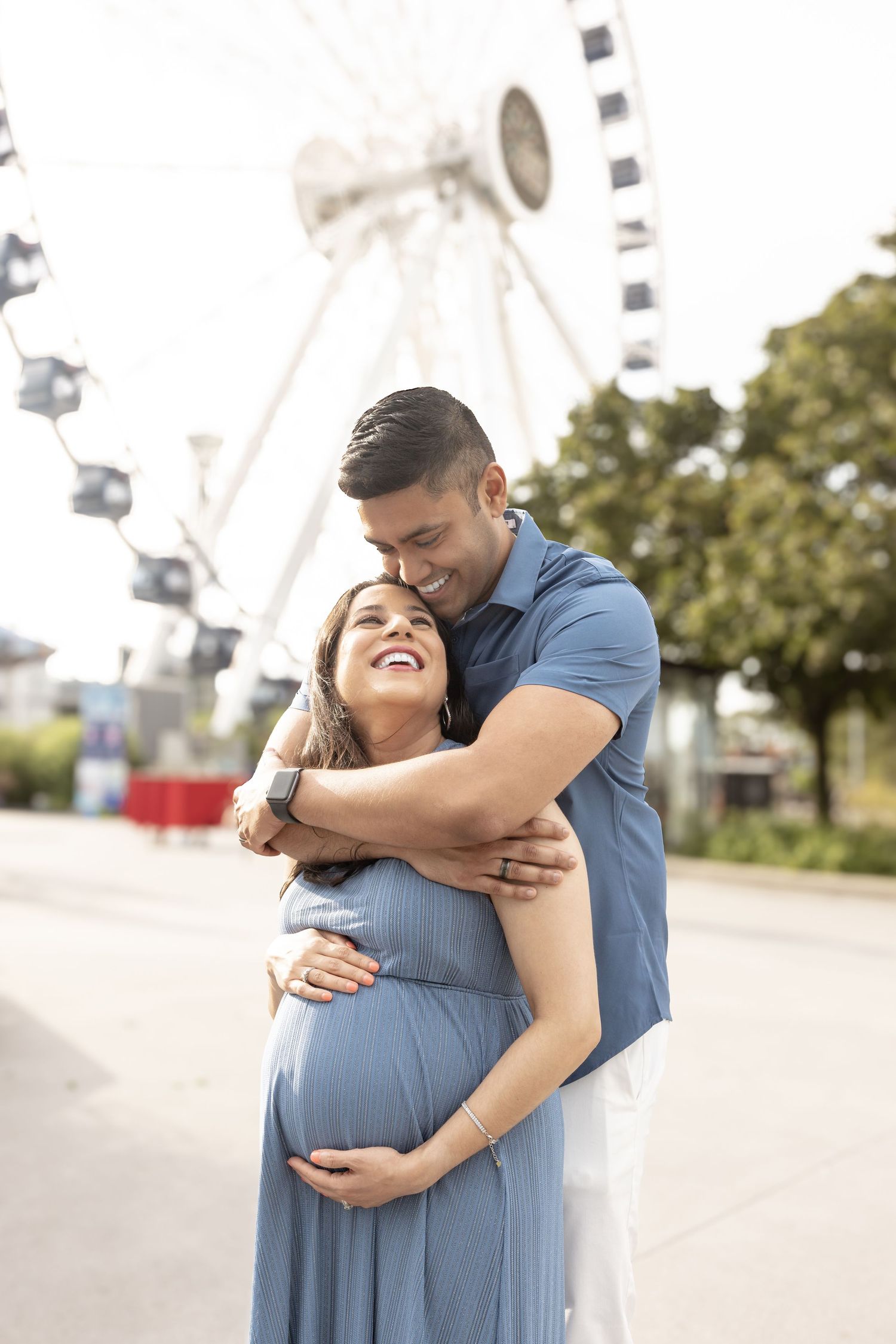 Man embracing pregnant woman from behind near Ferris wheel.