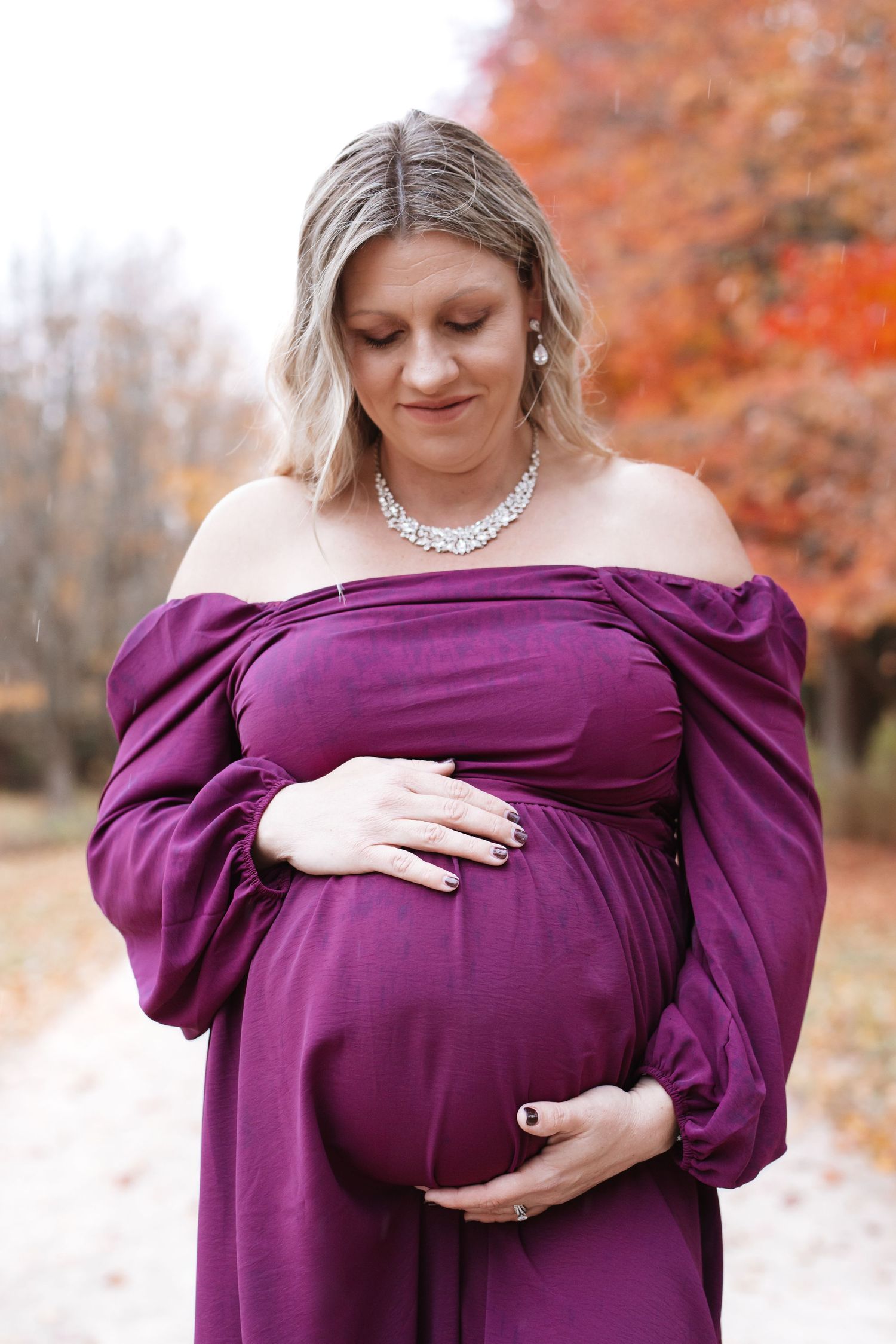 Pregnant woman in purple off-shoulder gown cradling baby bump outdoors amid vibrant autumn foliage.