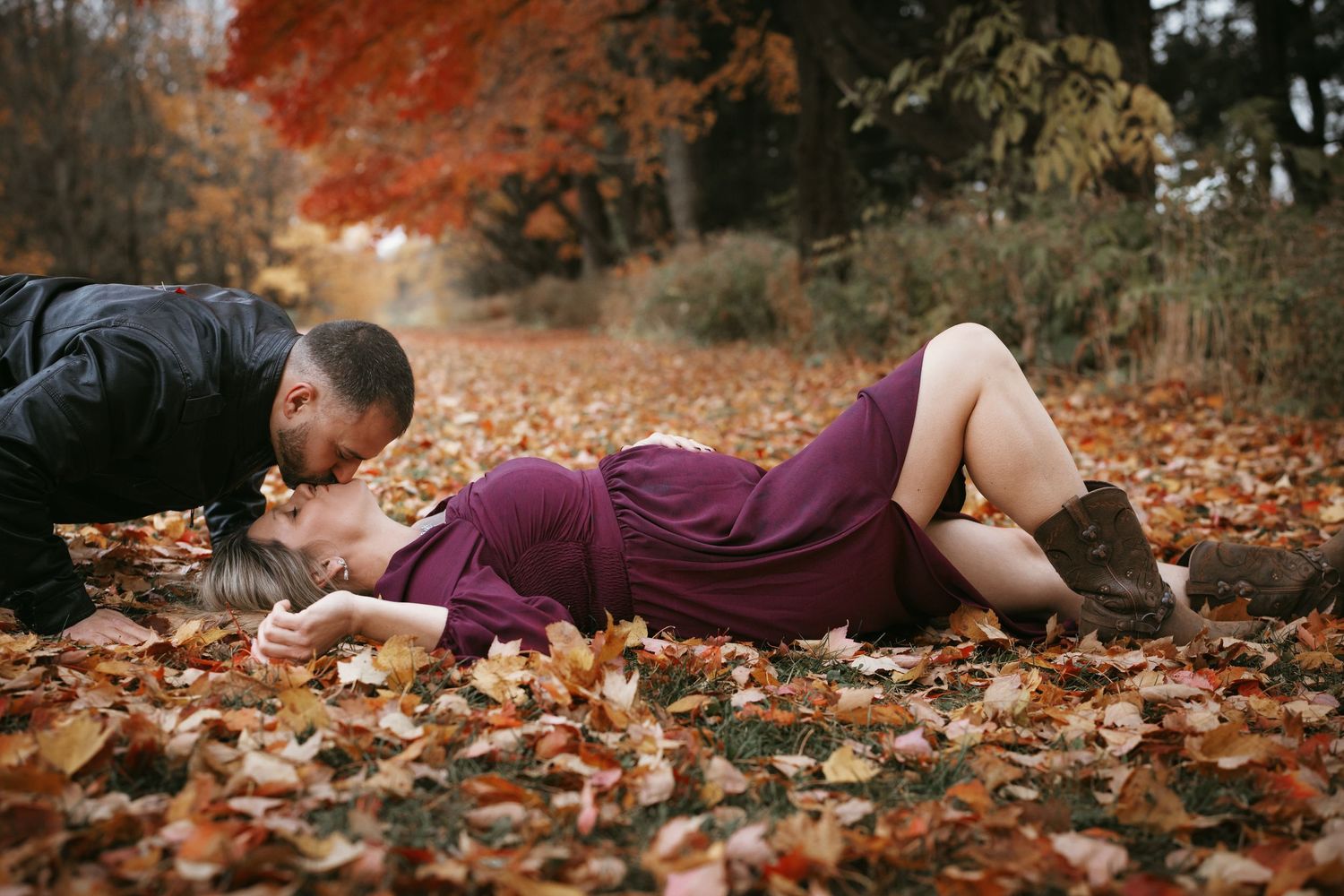 Man kissing pregnant woman lying on a bed of fall leaves in a vibrant autumn forest setting.