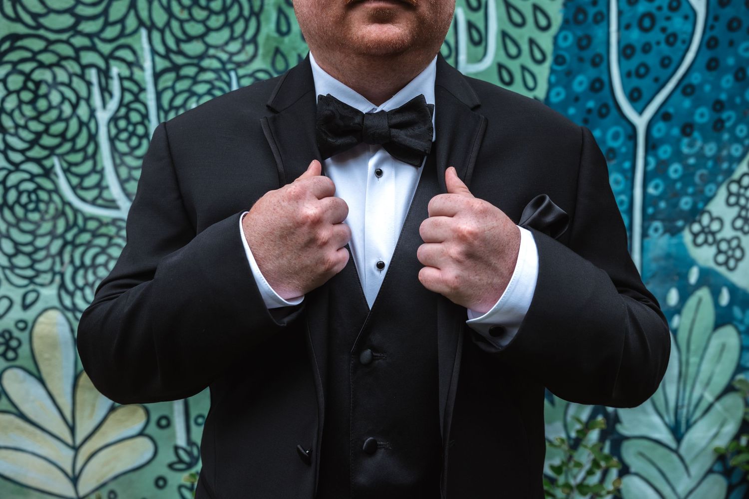 Close-up of groom in black tuxedo adjusting lapels with bow tie and pocket square visible.