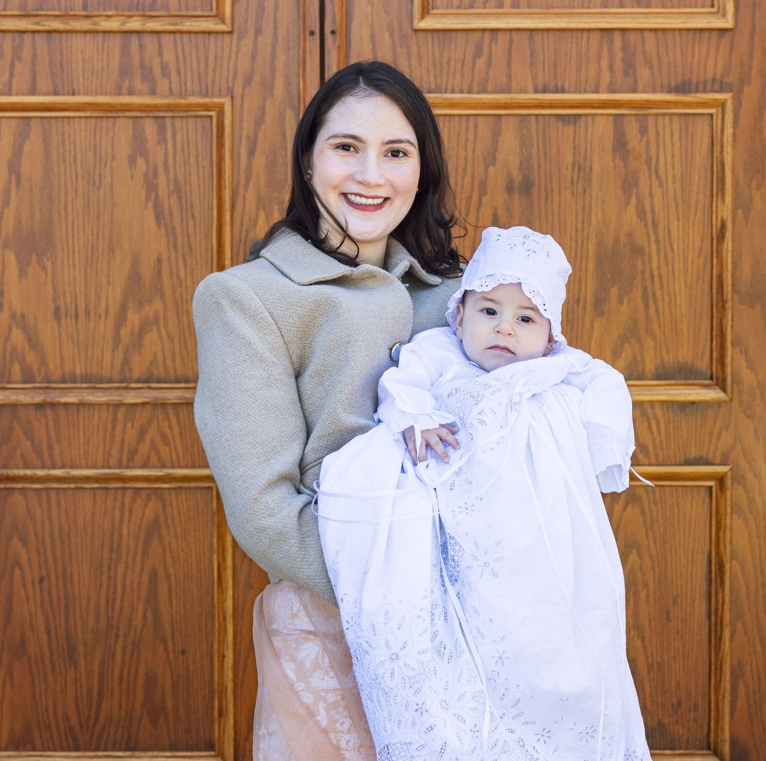 Young woman holds a baby dressed in a white christening gown and bonnet in front of wooden church doors.