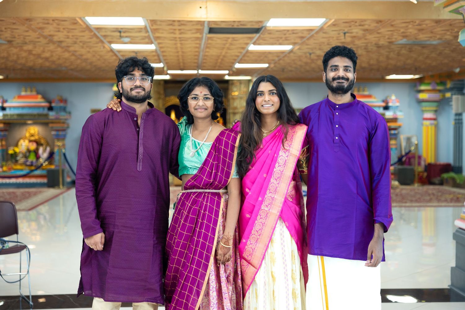 Four young adults in traditional Indian clothing posing together inside a vibrant Hindu temple.