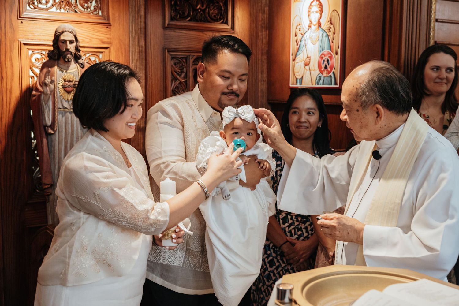 A Catholic priest anoints a baby while Filipino parents in white barong attire assist during baptism.