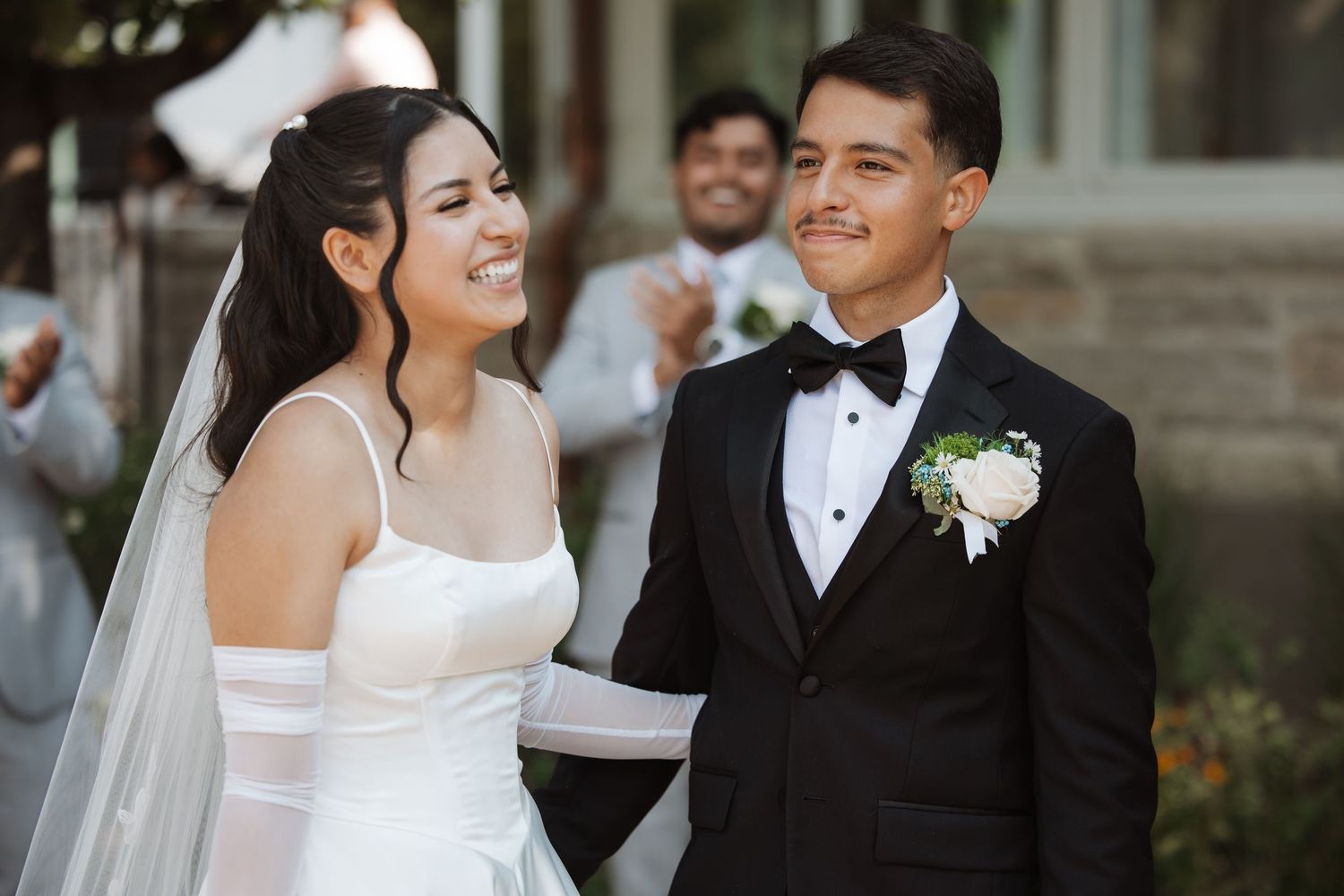 Smiling bride in white gown and groom in black tuxedo share joyful moment during wedding ceremony.