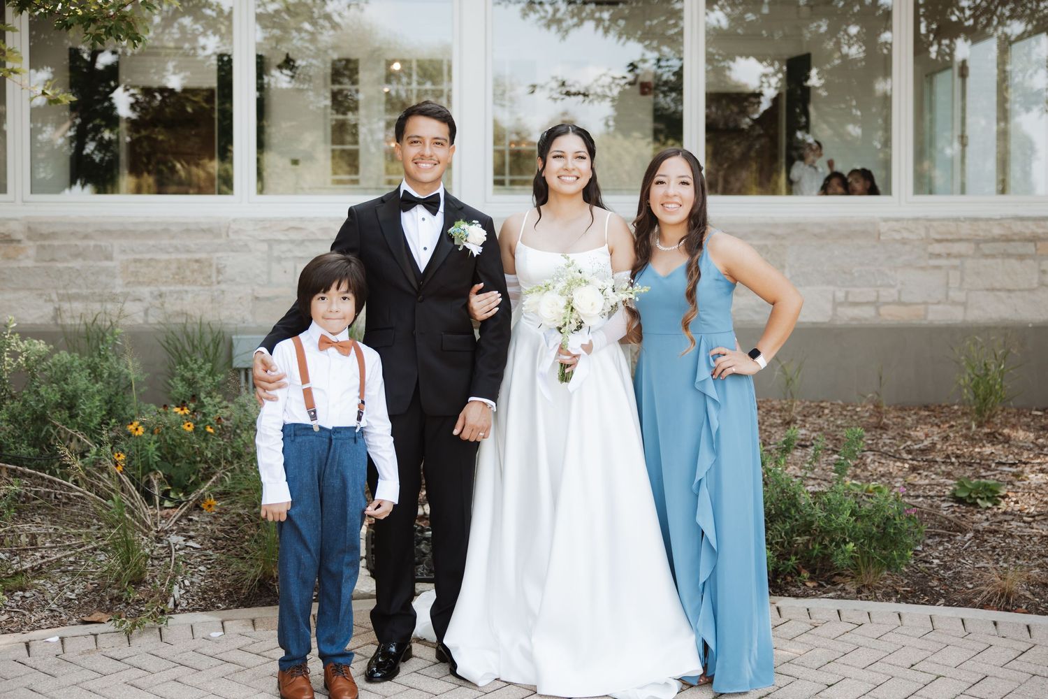 Groom in tuxedo and bride in white gown pose with young ring bearer and bridesmaid in blue dress.