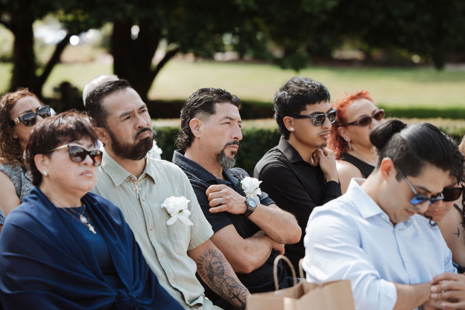 Group of wedding guests seated outdoors, attentively watching a wedding ceremony on a sunny day.