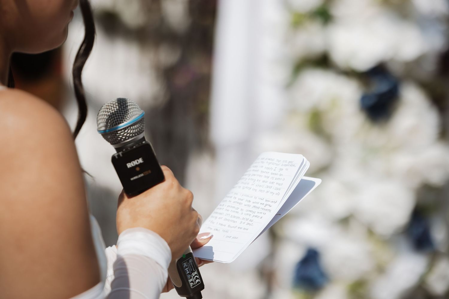 Bride reads wedding vows from a notebook while holding a RODE microphone at an outdoor ceremony.