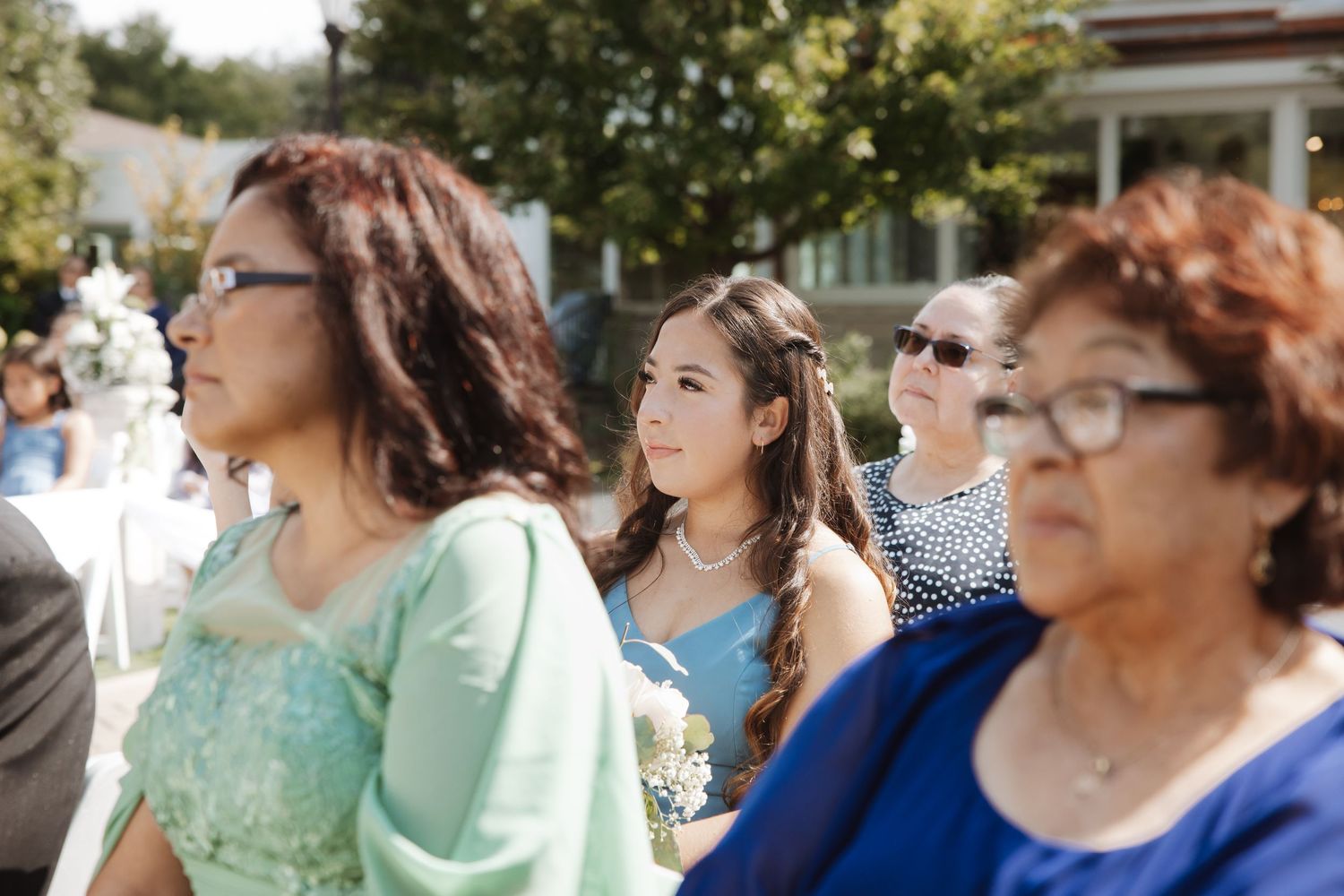 Wedding guests in colorful formal attire watch an outdoor ceremony on a bright sunny day.