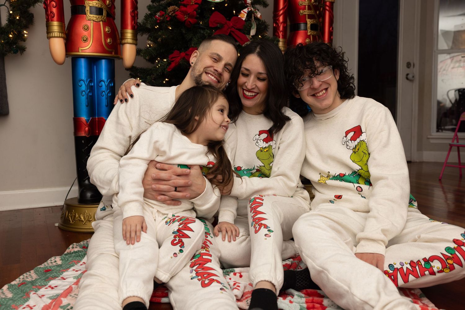 Joyful family of four in matching white Grinch Christmas pajamas cuddling by festive tree.