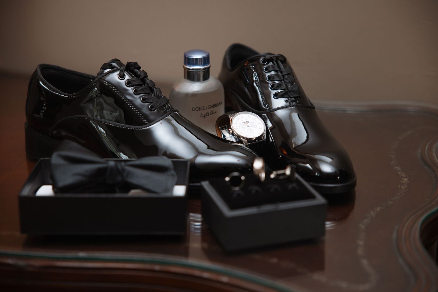 Elegant groom wedding accessories flat lay with patent leather shoes, cologne, watch, bow tie, and cufflinks in boxes.