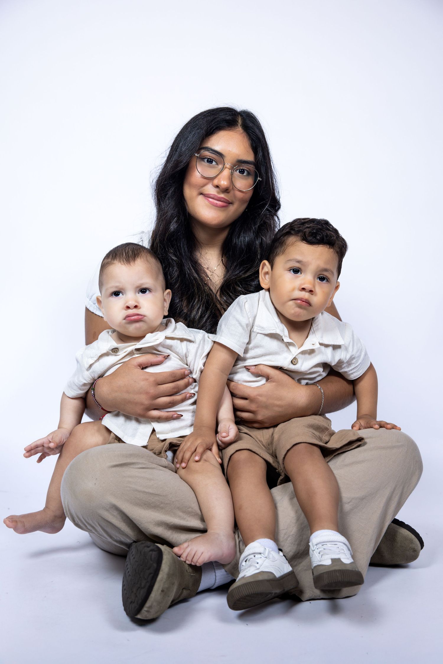 Young woman with glasses sitting on floor holding two young children in matching white and beige outfits.