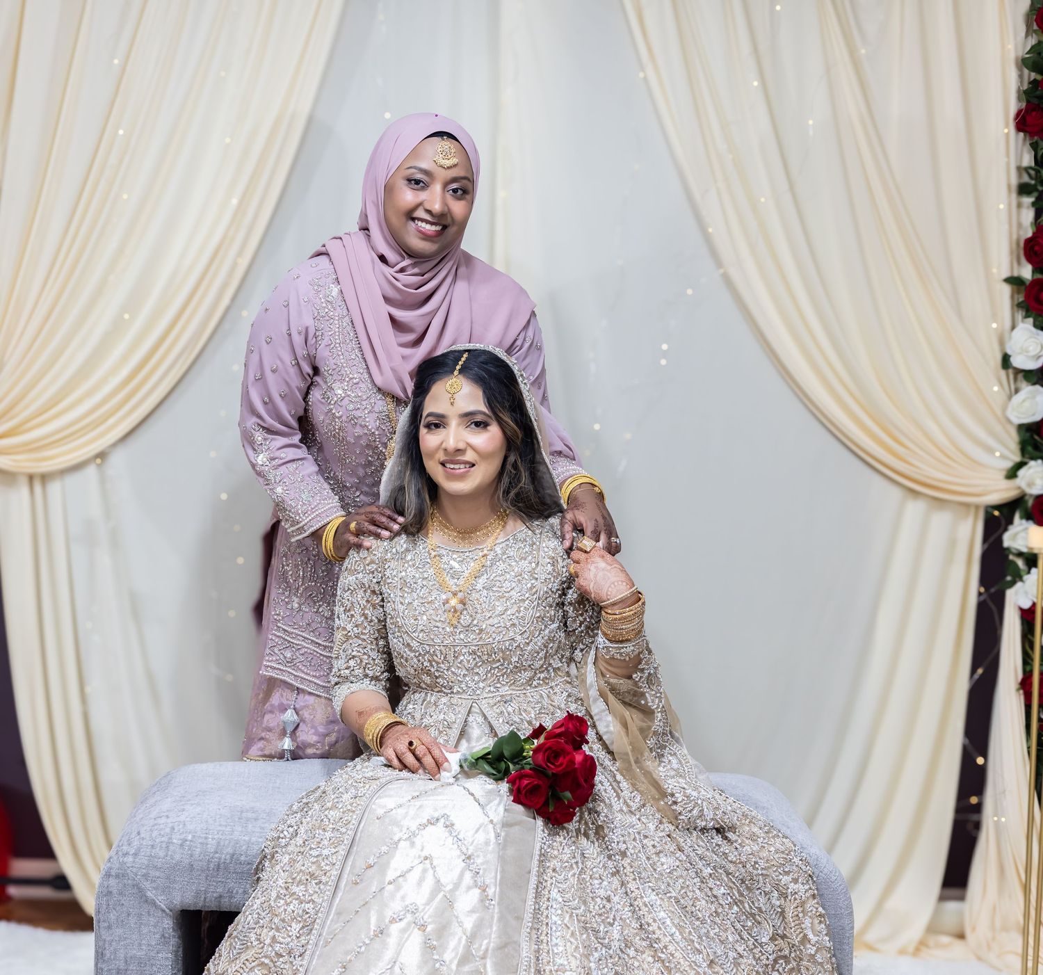 Bride in gold bridal lehenga holds red roses while woman in pink hijab stands beside at wedding backdrop.
