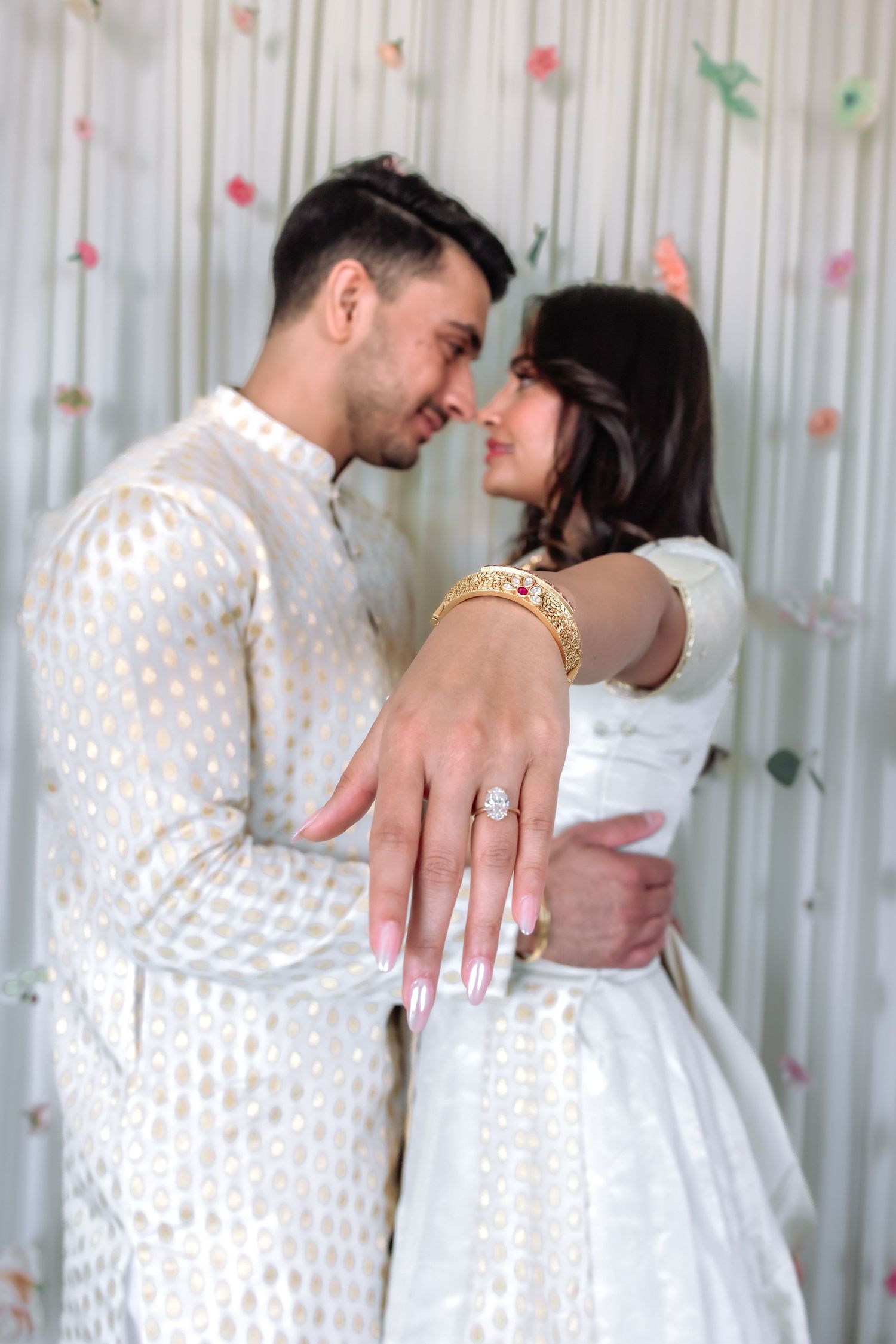 Indian couple in white traditional attire showing off diamond engagement ring at their engagement celebration.