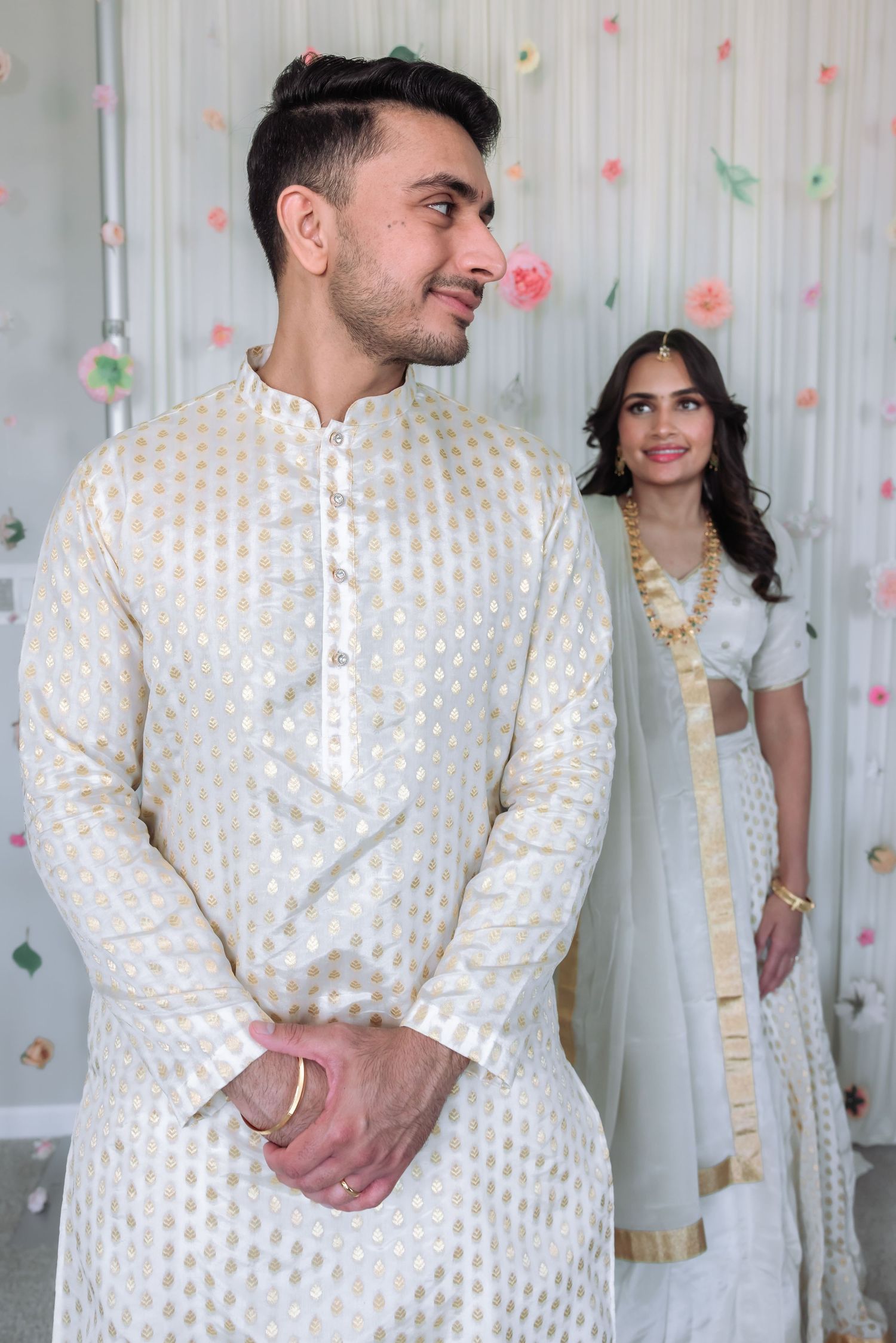 Young South Asian groom in white and gold kurta smiling with bride in background at traditional ceremony.