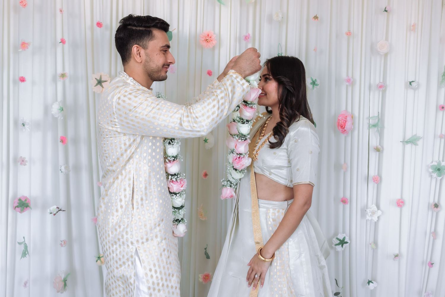 Indian groom places floral garland on bride during traditional Hindu engagement ceremony in white attire.