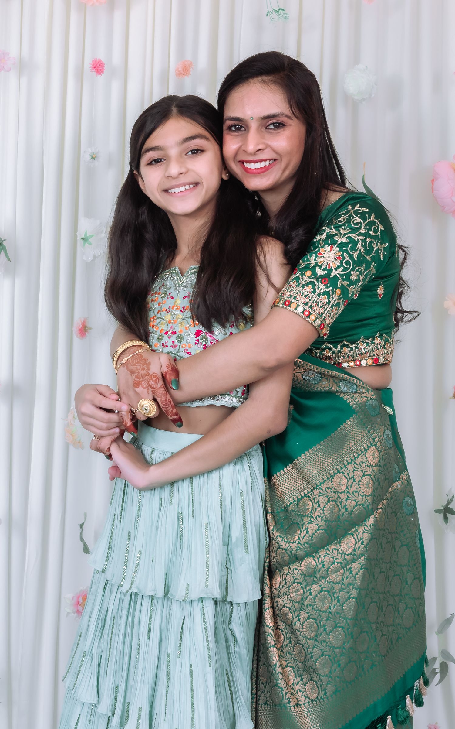 Woman in green saree hugs smiling girl in mint lehenga with mehndi at Indian celebration.