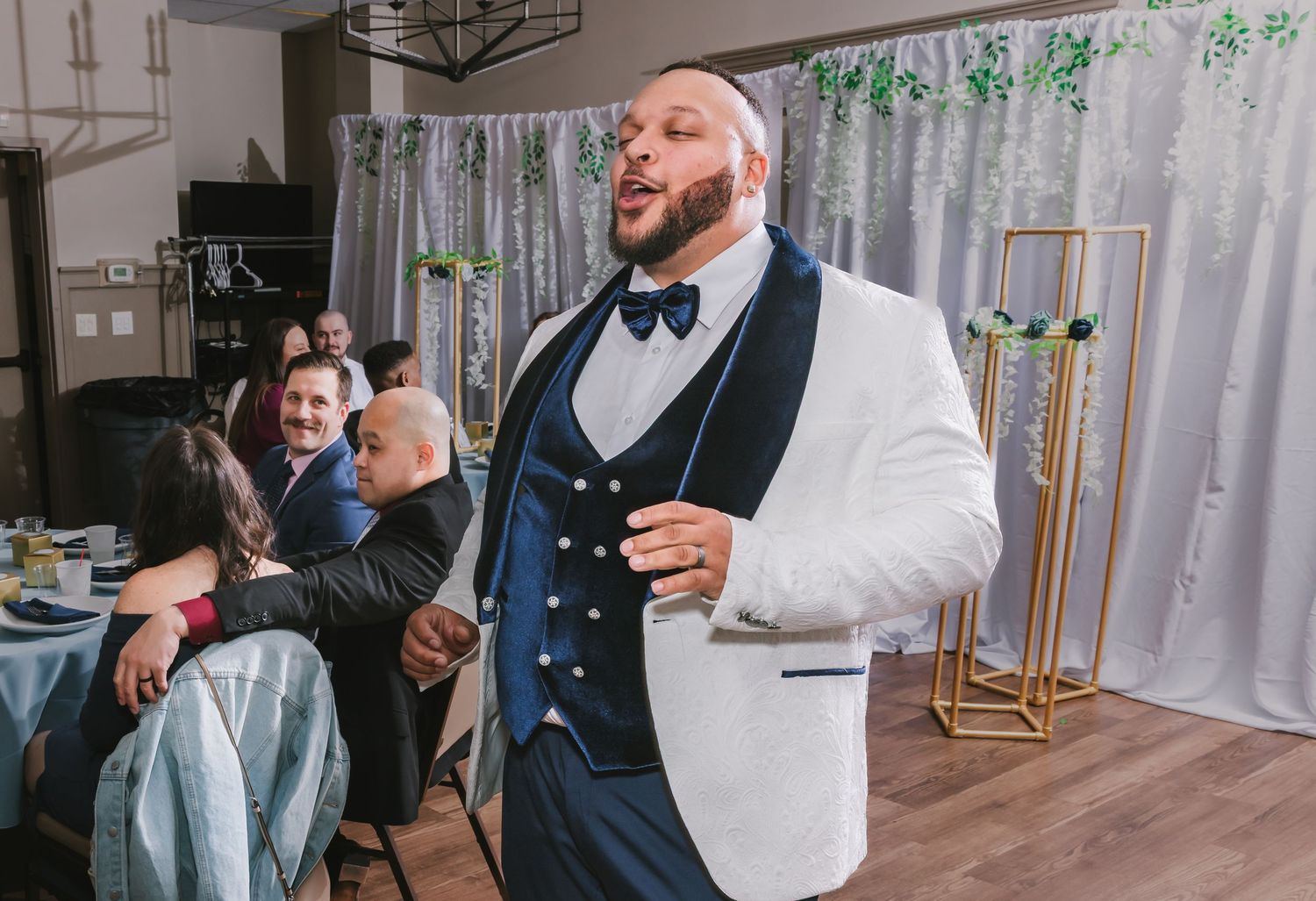 Groom in white tuxedo jacket and navy bow tie smiles at elegant wedding reception celebration.