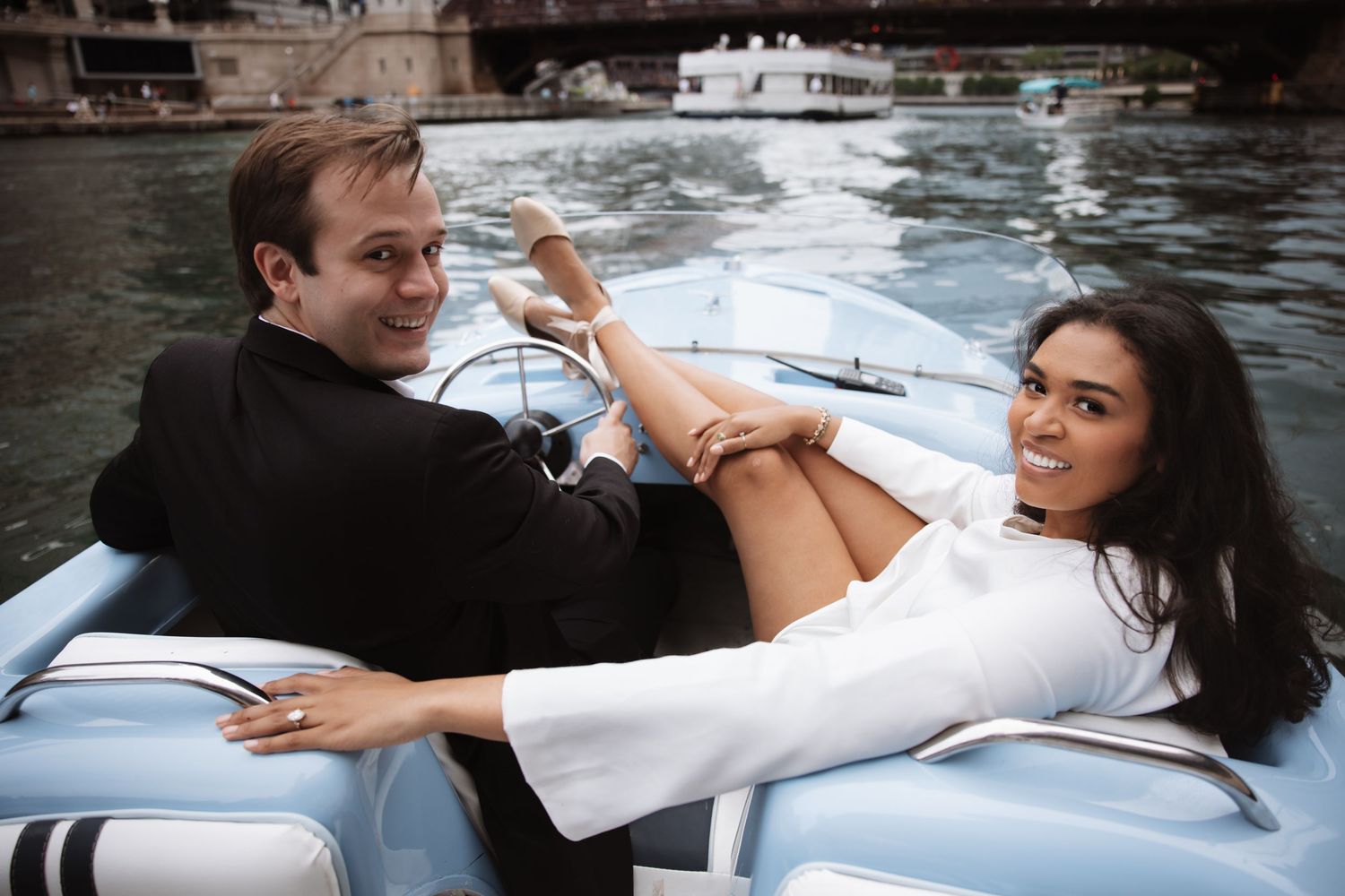 Couple smiling and relaxing on a classic blue boat on the river, woman in white dress reclining across man in black suit.