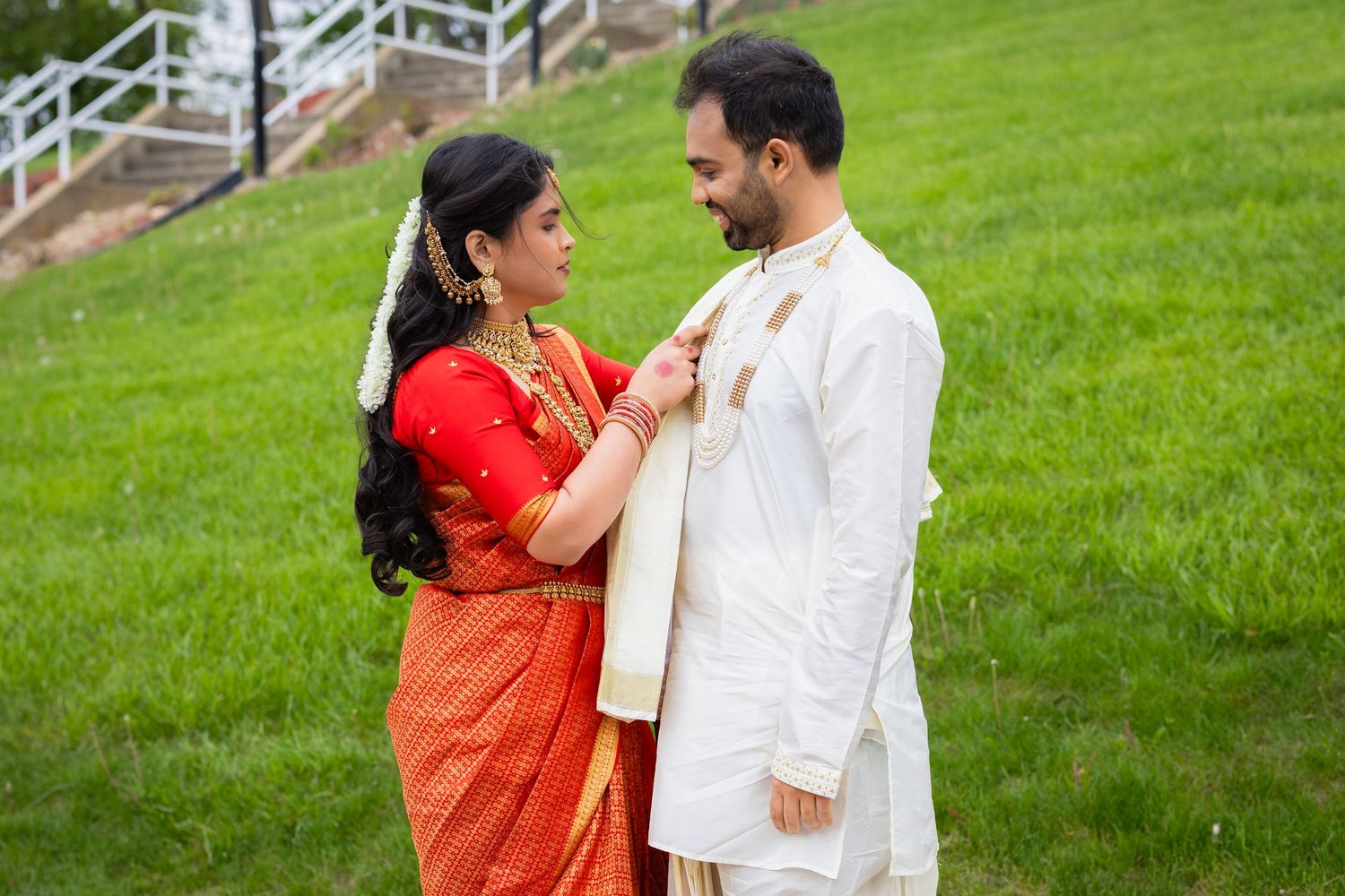 Indian couple in traditional wedding attire, bride in orange saree and groom in white sherwani, sharing a romantic moment outdoors.