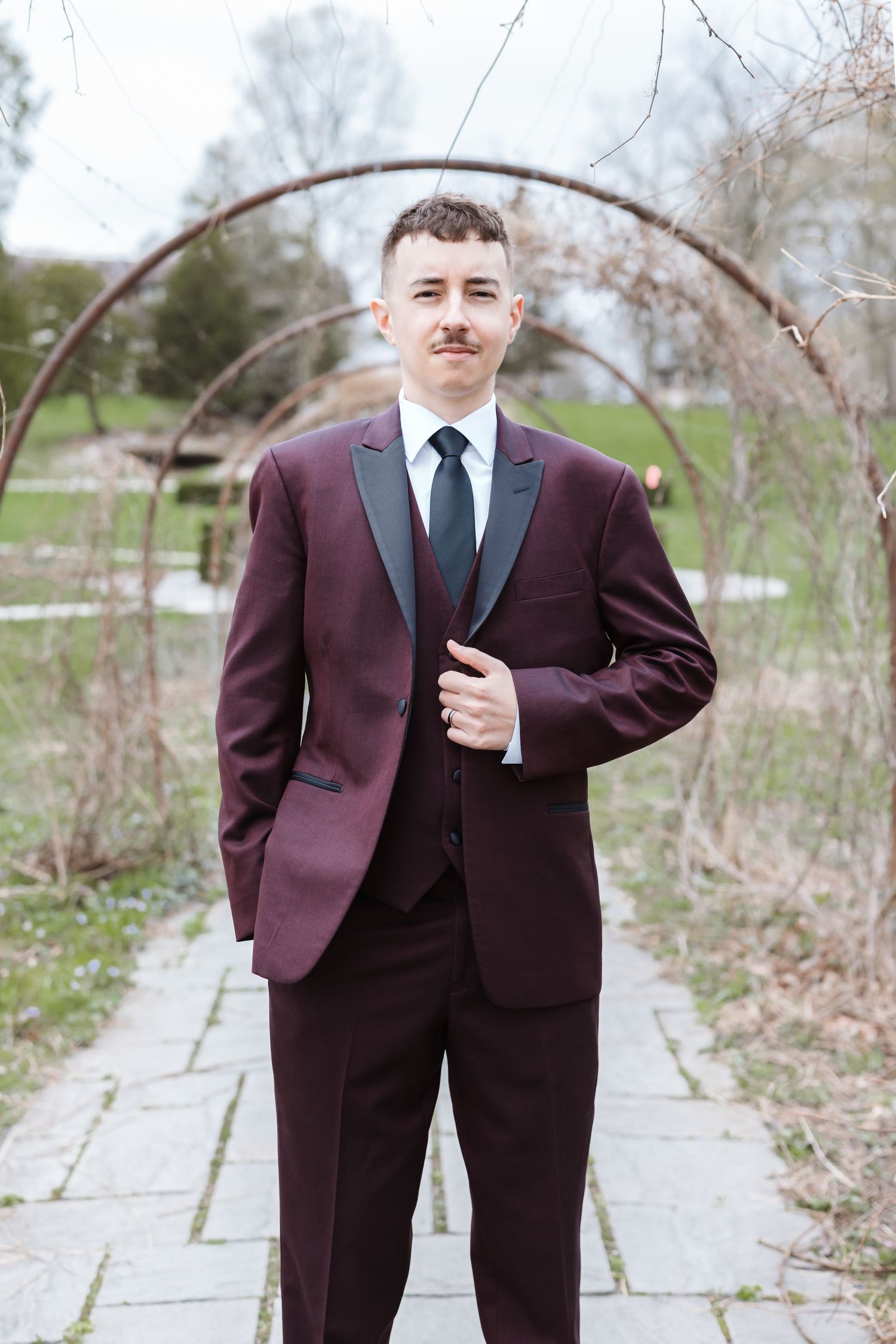 Young man in burgundy tuxedo with black lapels and tie poses on stone path under rustic arch in outdoor garden setting.