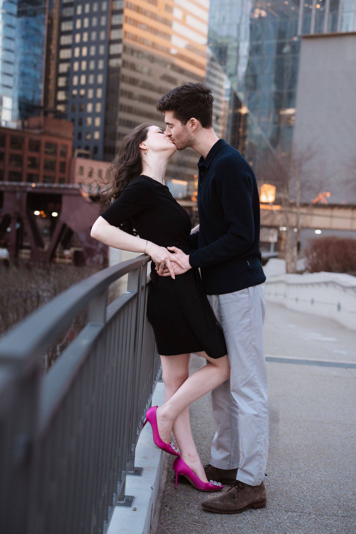 A couple kisses while holding hands on a urban bridge, woman wearing pink heels and black dress.