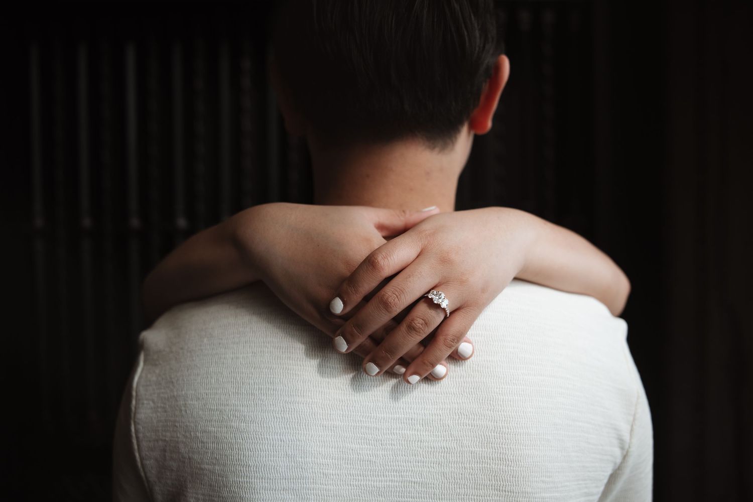 Close-up of woman's hand with diamond engagement ring wrapped around man's neck in tender embrace.