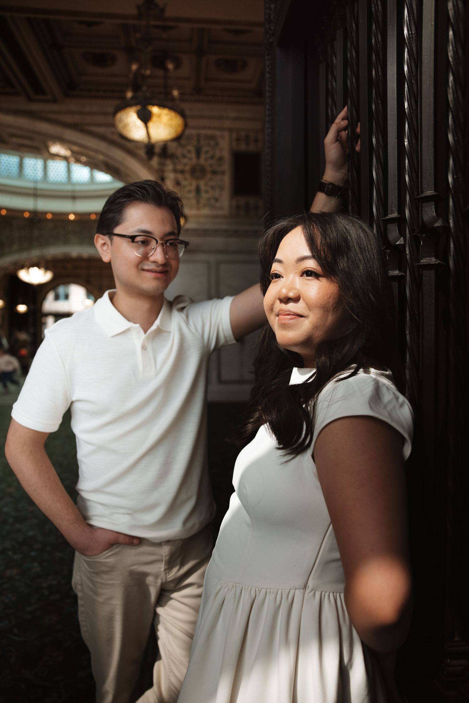 Couple posing by ornate black iron gate in elegant historic building, woman in white dress smiling warmly.