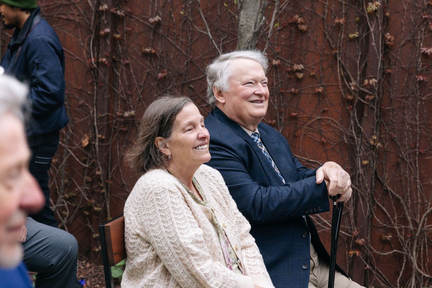 An older couple smiles joyfully while seated outdoors at a wedding reception near a rustic vine-covered wall.