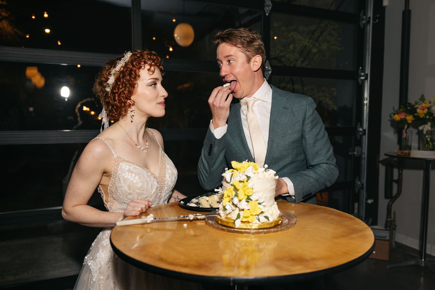 Bride feeds groom wedding cake at candlelit table with yellow floral bouquet during reception.