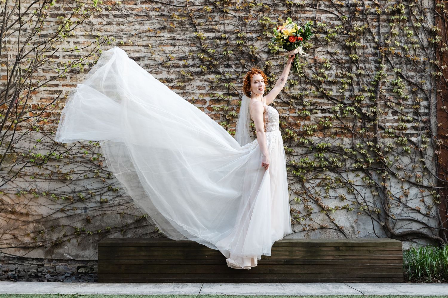 Joyful red-haired bride twirls her flowing white wedding gown while raising her colorful bouquet against a brick wall.
