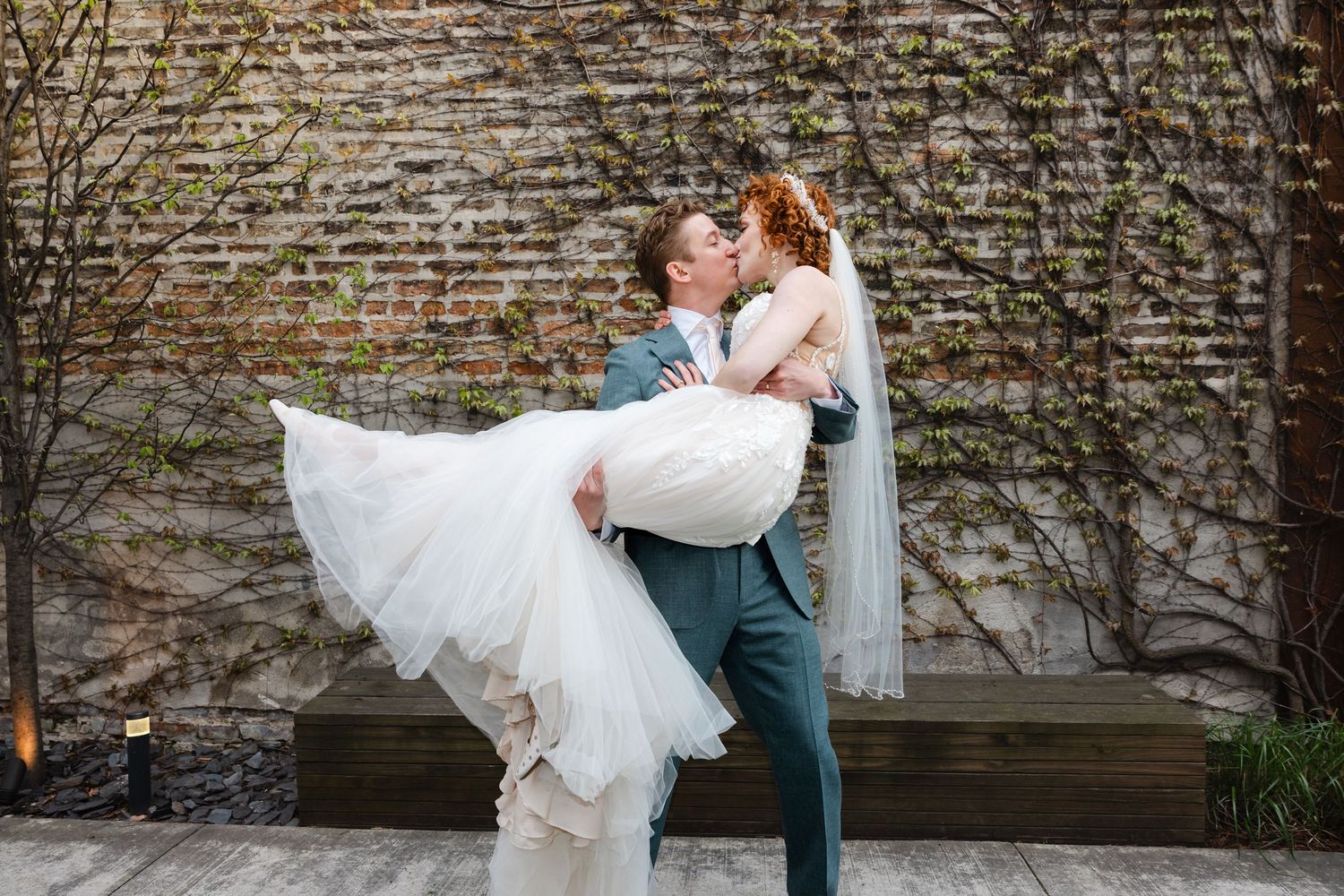 Groom lifts and kisses bride in flowing white wedding dress outdoors against ivy-covered brick wall backdrop.