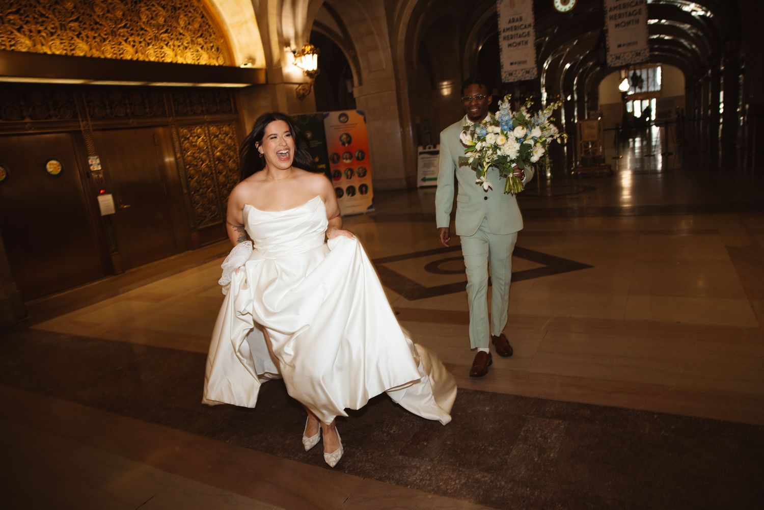 Bride in white gown and groom in mint suit walk through ornate venue hall, carrying a lush floral bouquet.