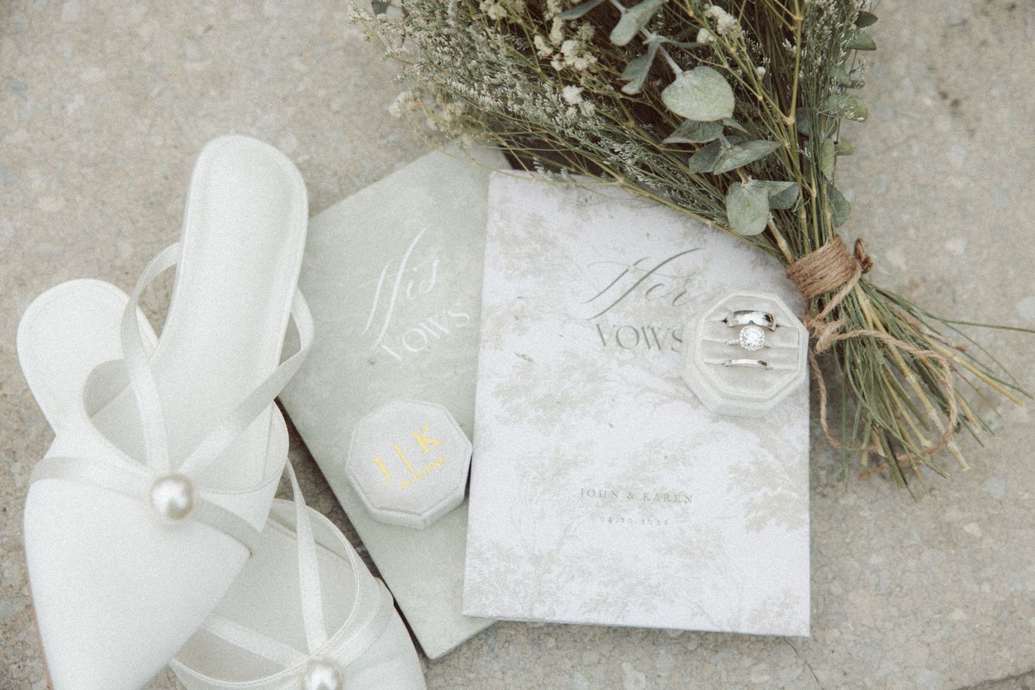 Elegant wedding flat lay with white heels, vow books, rings, candle, and eucalyptus bouquet on gray surface.